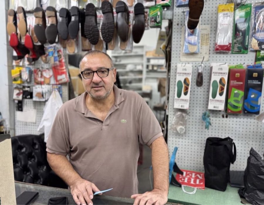 Arsen Khochaba, founder of AK Shoe Repair, standing at the shop's repair counter with rows of soles and heels on the wall behind him