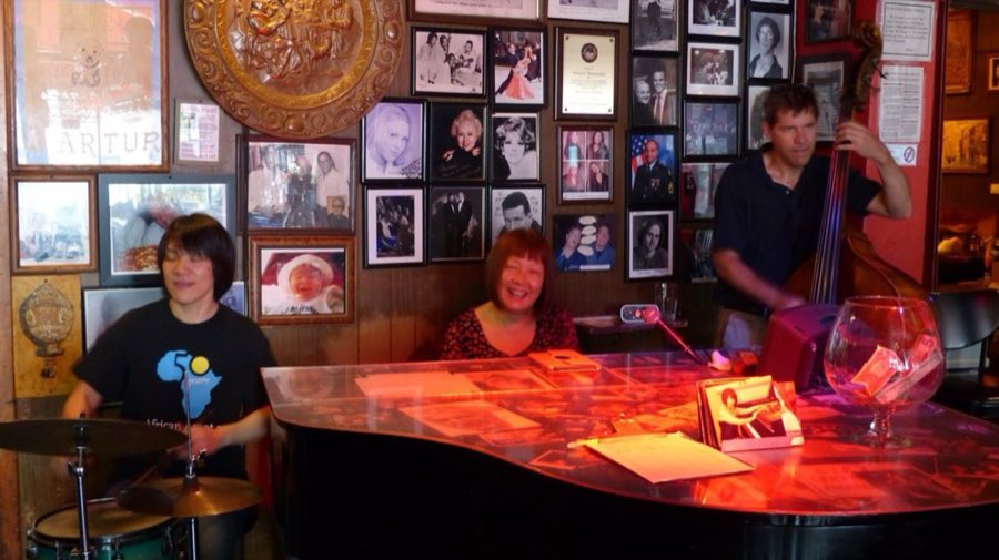 A jazz trio — drummer, pianist and upright bassist — performing under warm red light in front of a wall densely covered with black-and-white photographs and plaques