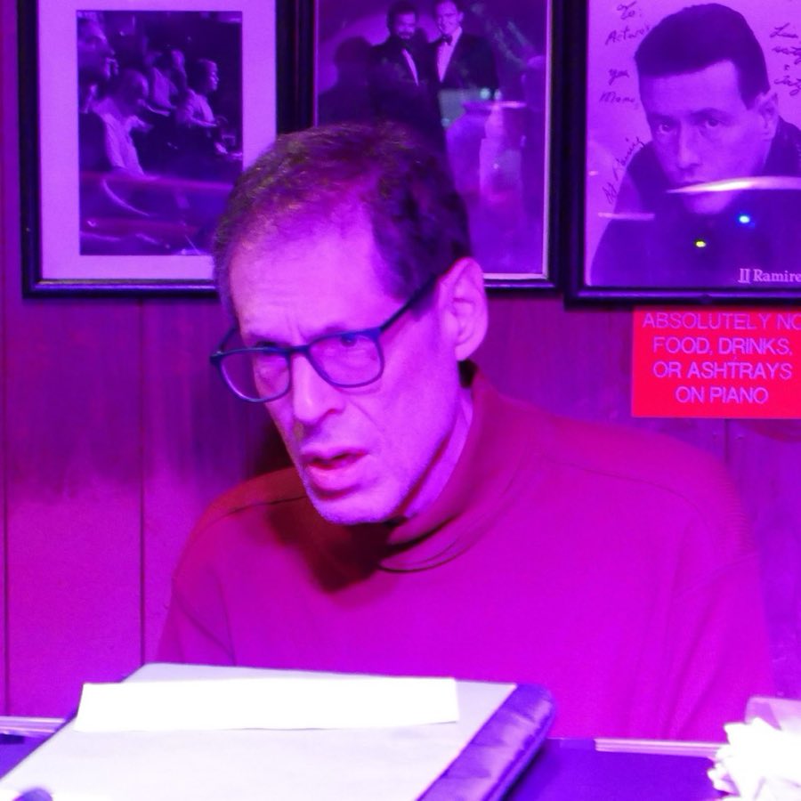 A musician at the piano under purple stage lighting, with a photo-covered wall behind