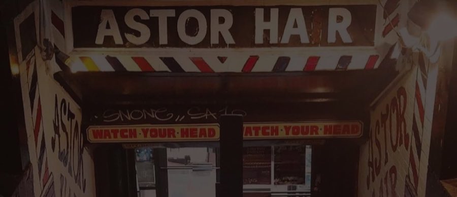 The barber-pole-striped ASTOR HAIR sign glowing over the basement stairwell entrance at 2 Astor Place, with a 'Watch Your Head' warning sign below it