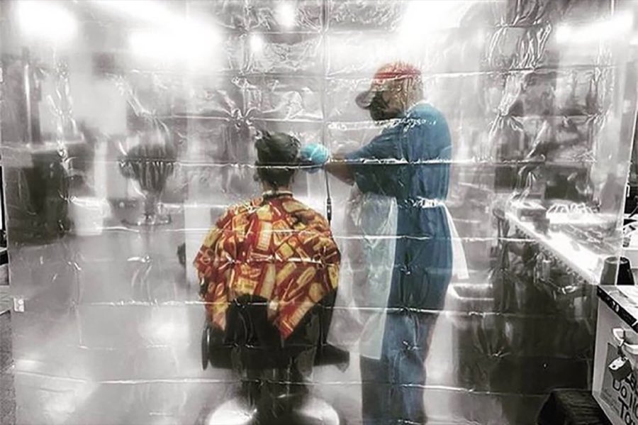 A barber in full protective gear cutting a customer's hair behind sheets of plastic sheeting hung floor to ceiling as a pandemic safety barrier