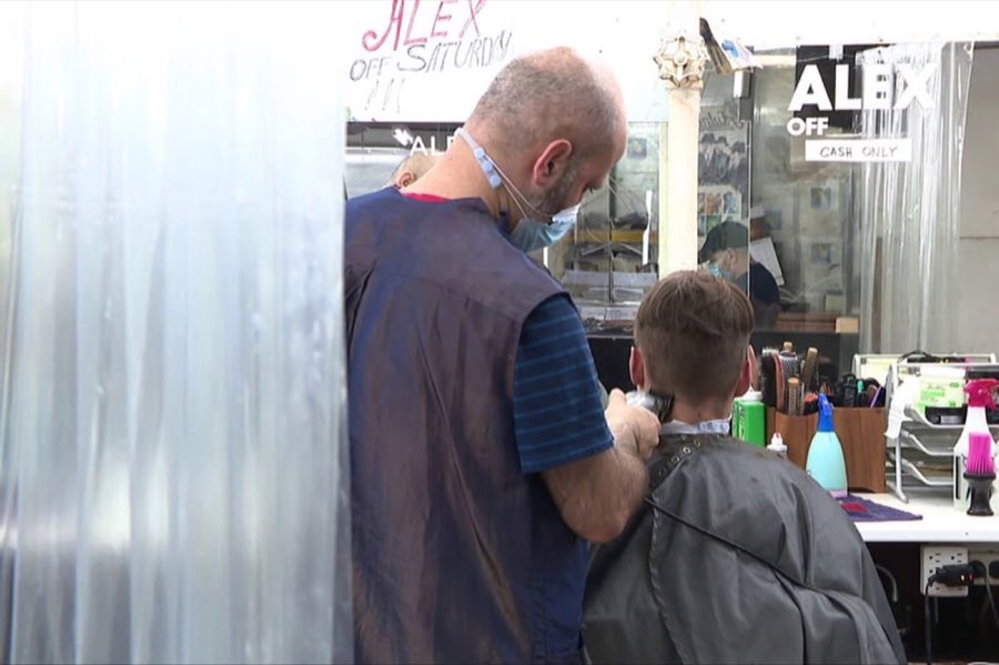 A barber cutting a boy's hair through translucent plastic sheeting hung as a pandemic-era safety barrier