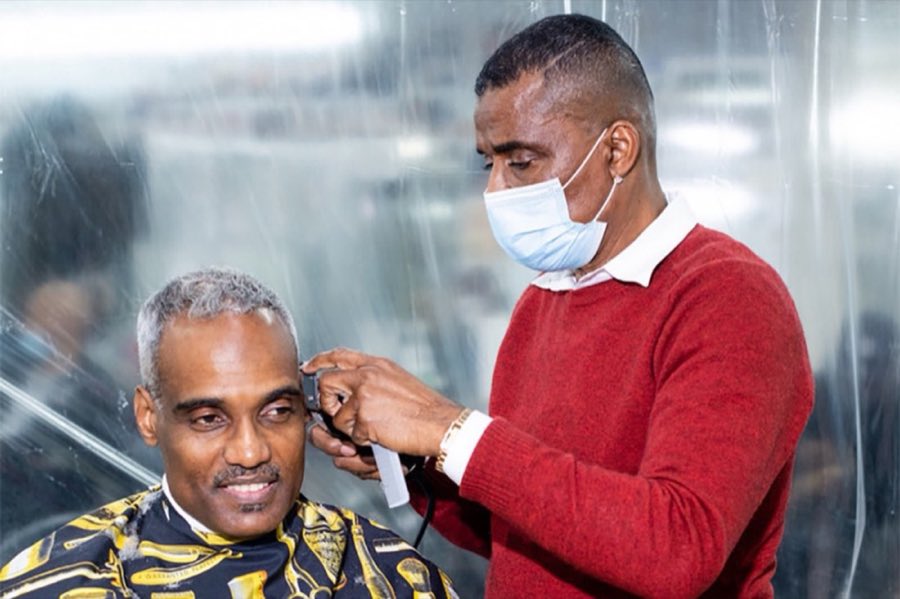 A masked barber giving a customer a head shave, the customer wearing a gold and black patterned barber cape