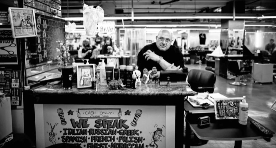 Black-and-white portrait of Mike Saviello seated at the shop's front desk, surrounded by taped-up photos, the hand-painted 'We Speak' sign, and a 'Cash Only, Thank You' notice