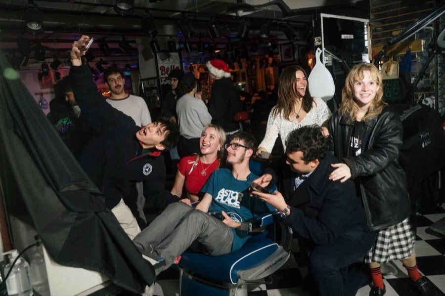 A lively nighttime party inside the shop with young customers laughing in barber chairs, one wearing an Astor Place: The American Dream t-shirt, on the black-and-white checkerboard floor