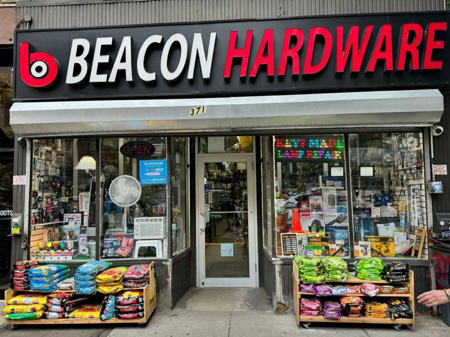 Beacon Hardware storefront at 371 Amsterdam Avenue: black fascia sign with white BEACON and red HARDWARE lettering, a red circular b logo, a lit OPEN sign, a Keys Made / Lamp Repair window sign, and bags of garden soil on sidewalk carts out front