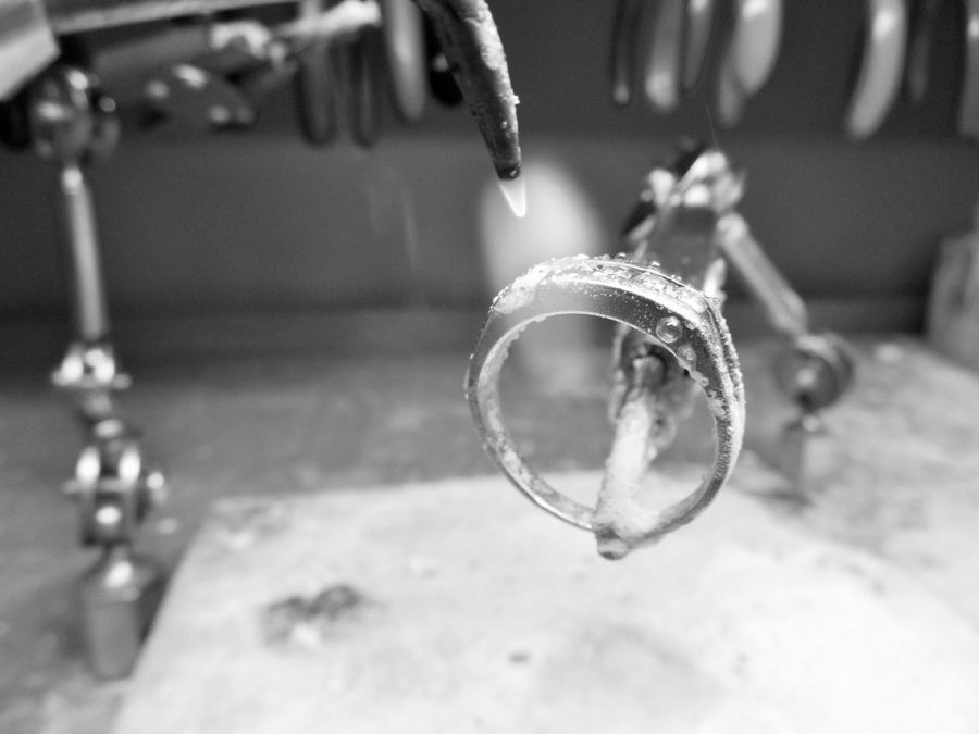 A ring held in tweezers beneath a jeweler's torch flame, mid-repair