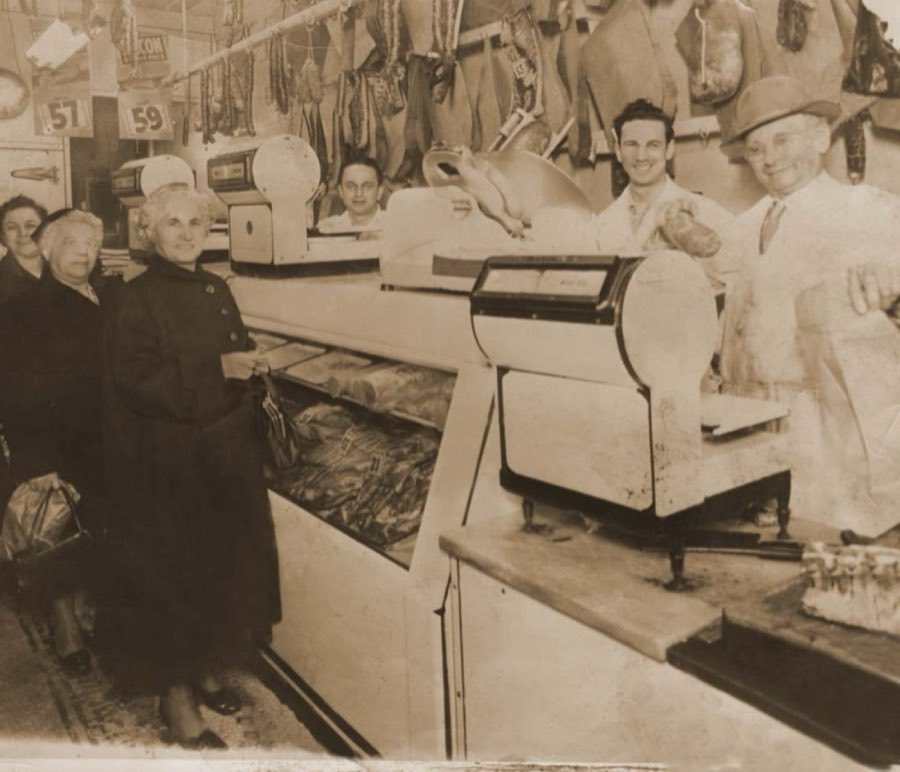 Sepia photograph of the shop interior in an earlier era, showing an old counter scale, hanging cured sausages, two customers, and three staff behind the counter