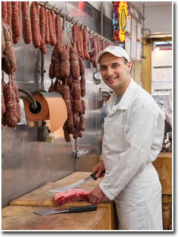 Robert Esposito smiling behind the counter, knife in hand next to a cut of meat, with hanging sausages and a Boar's Head sign behind him