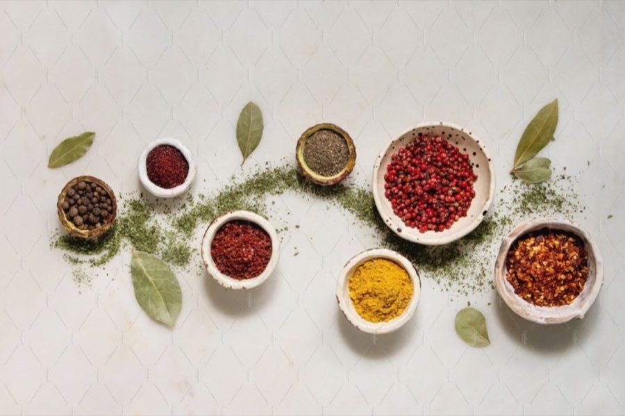 Overhead flat-lay of sumac, black and pink peppercorns, chili flakes, turmeric and bay leaf in small bowls on a marble surface