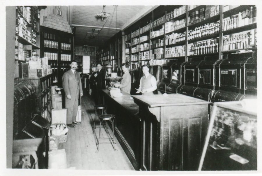 Inside the early Katagiri store: a long wooden counter with staff in shirtsleeves, shelves stacked with tinned goods, and a painted "Teas & Coffees" sign.