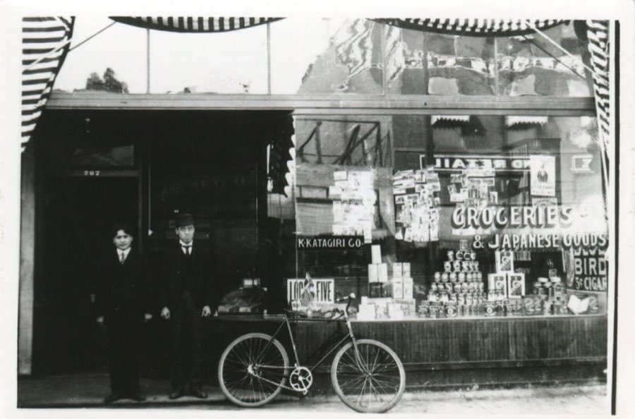 The original K. Katagiri Co. storefront in the early 1900s: two young men in dark suits and a bicycle outside a shop window hand-painted "Groceries & Japanese Goods".