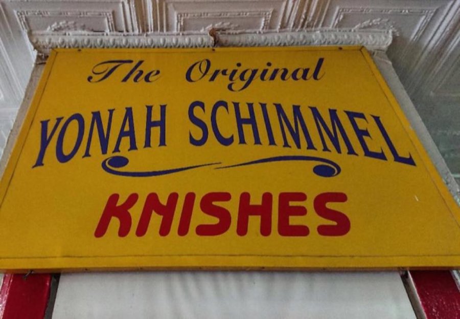 The hand-painted Yonah Schimmel Knishes storefront sign — yellow board with navy script lettering and bold red 'KNISHES' text, mounted beneath an ornate pressed-tin ceiling