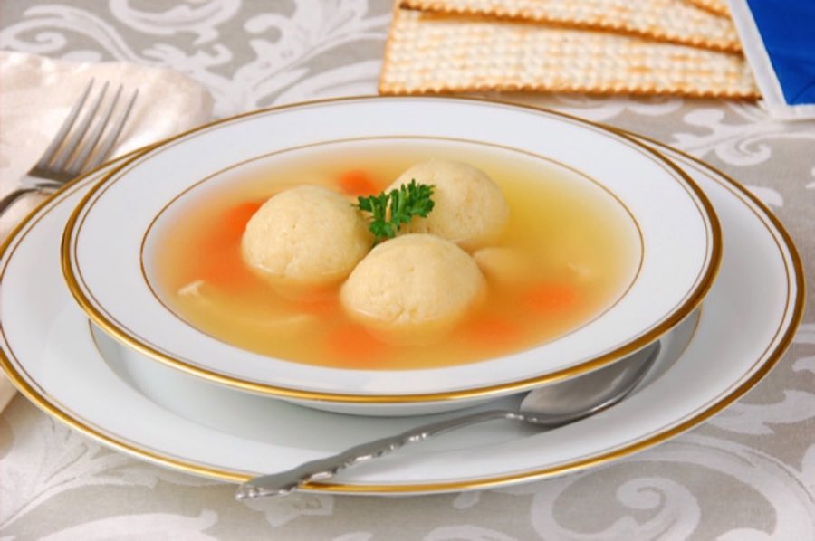 Three matzo balls in golden chicken broth with carrots and parsley, in a gold-rimmed bowl beside matzo