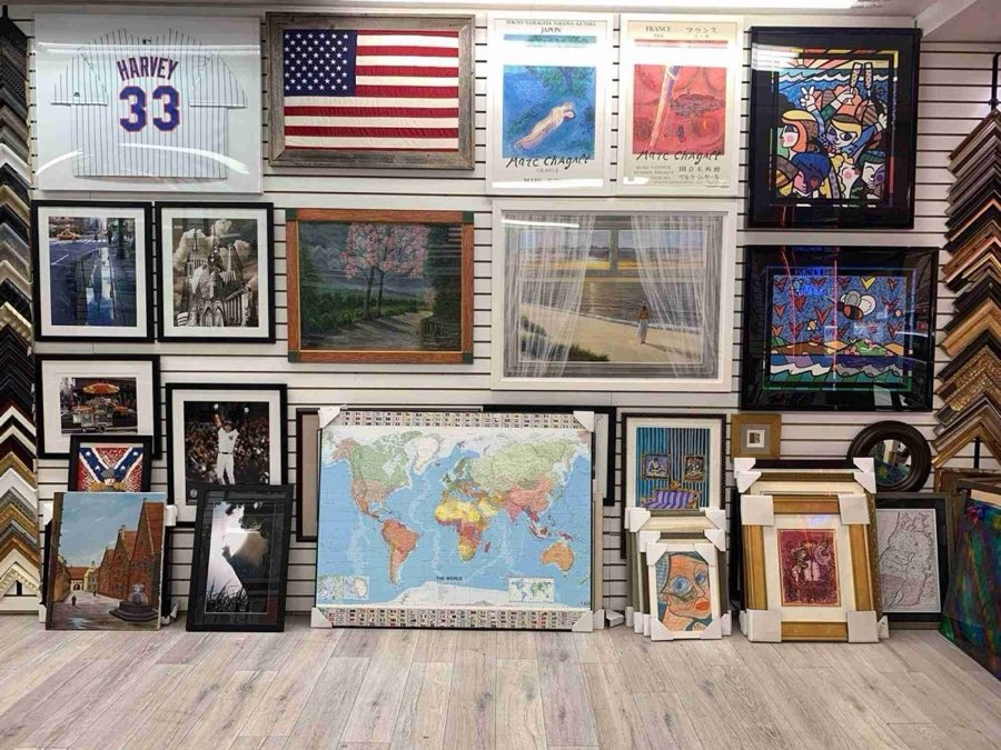 Interior wall of the shop covered with framed pieces: a Mets jersey, an American flag, Marc Chagall museum posters, colorful pop-art prints, black-and-white New York City photographs, a landscape painting, and a world map, with unfinished frames leaning against the herringbone wood floor