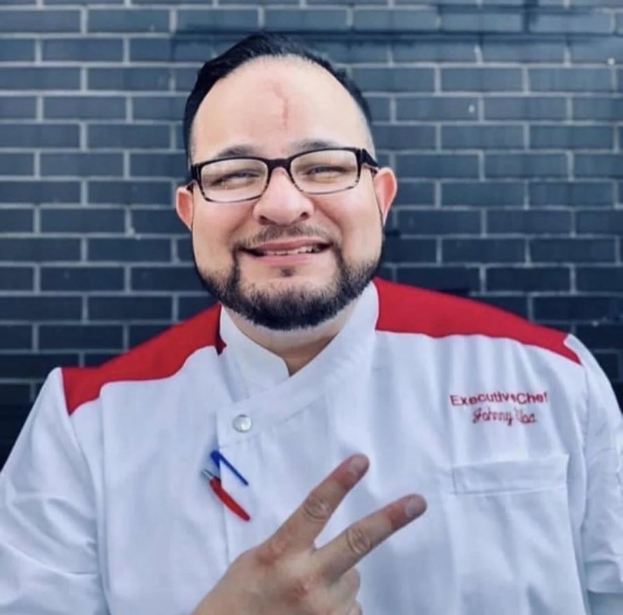Executive chef Johnny Ulloa in an embroidered white chef coat, smiling against a dark brick wall
