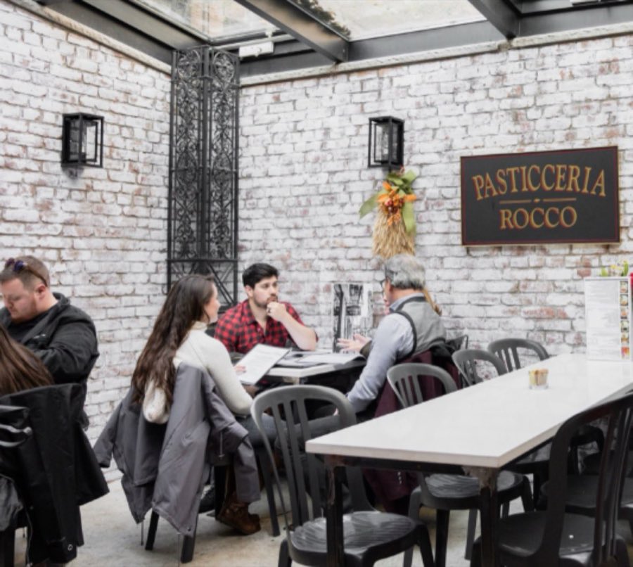 Guests seated at a café table in Rocco&rsquo;s whitewashed-brick garden room, beneath the black-and-gold Pasticceria Rocco sign