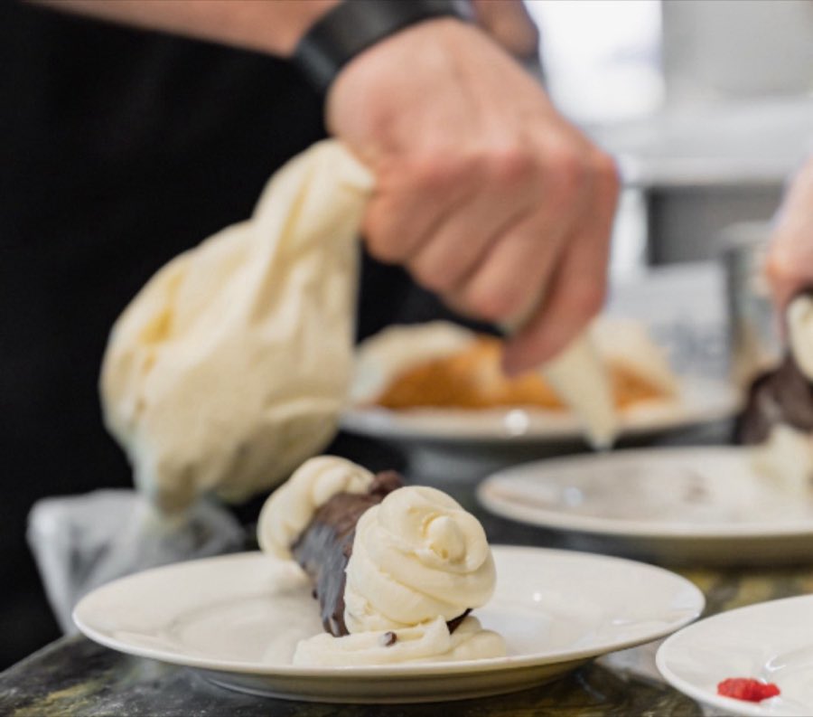 A baker&rsquo;s hands piping ricotta cream into a chocolate cannoli shell on a plate