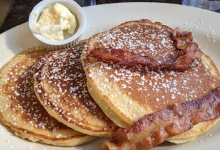 A breakfast plate of golden pancakes with bacon, powdered sugar and a ramekin of butter