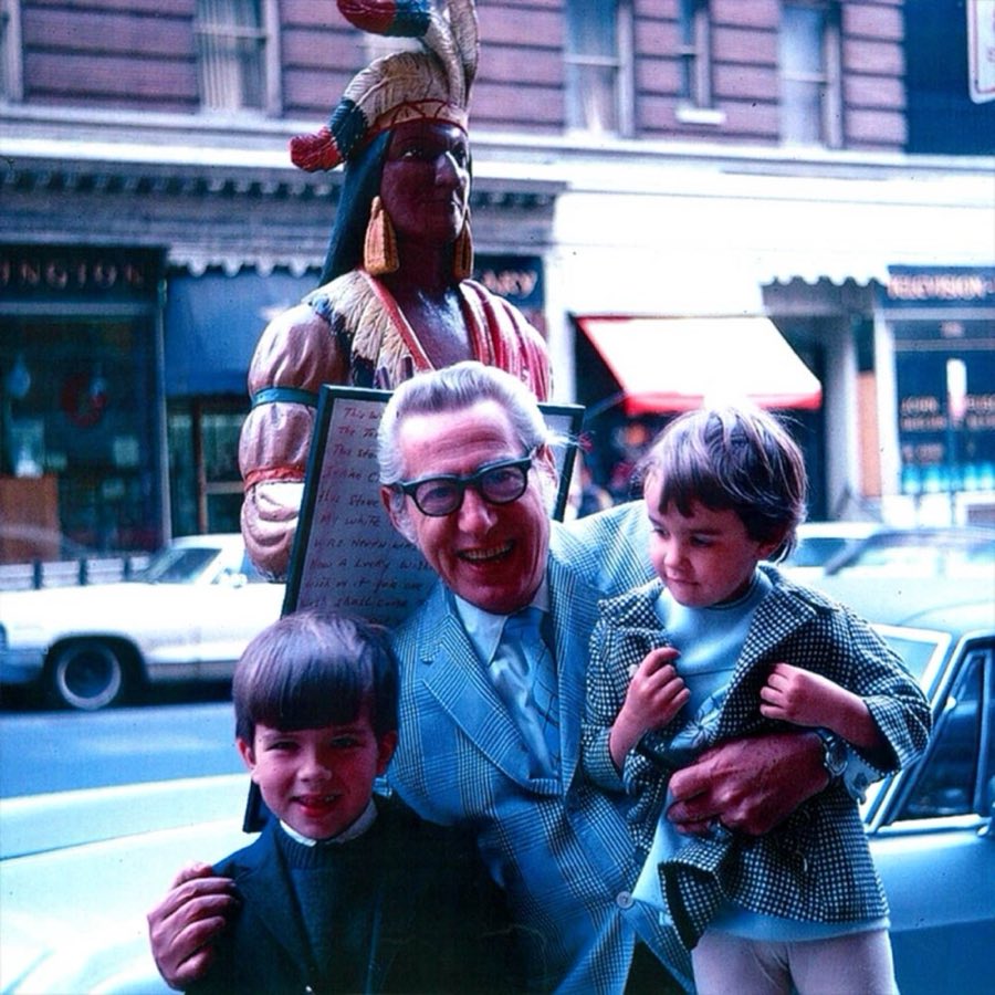 A vintage color photograph of an older man with two young boys standing on the Lexington Avenue sidewalk in front of the shop's carved wooden trade figure
