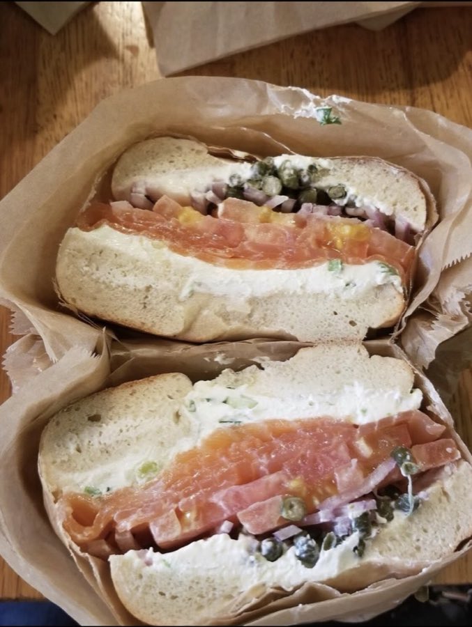 A lox bagel sandwich sliced in half, piled with cream cheese, smoked salmon, capers, tomato and red onion, wrapped in deli paper