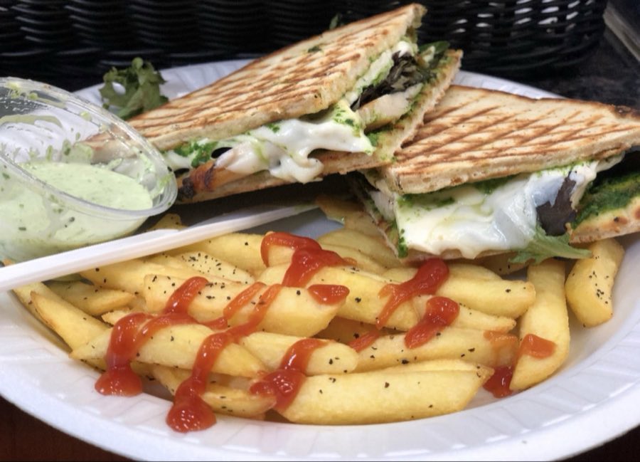 A grilled panini with melted cheese and pesto, cut diagonally, served with french fries and ketchup and a cup of green herb sauce