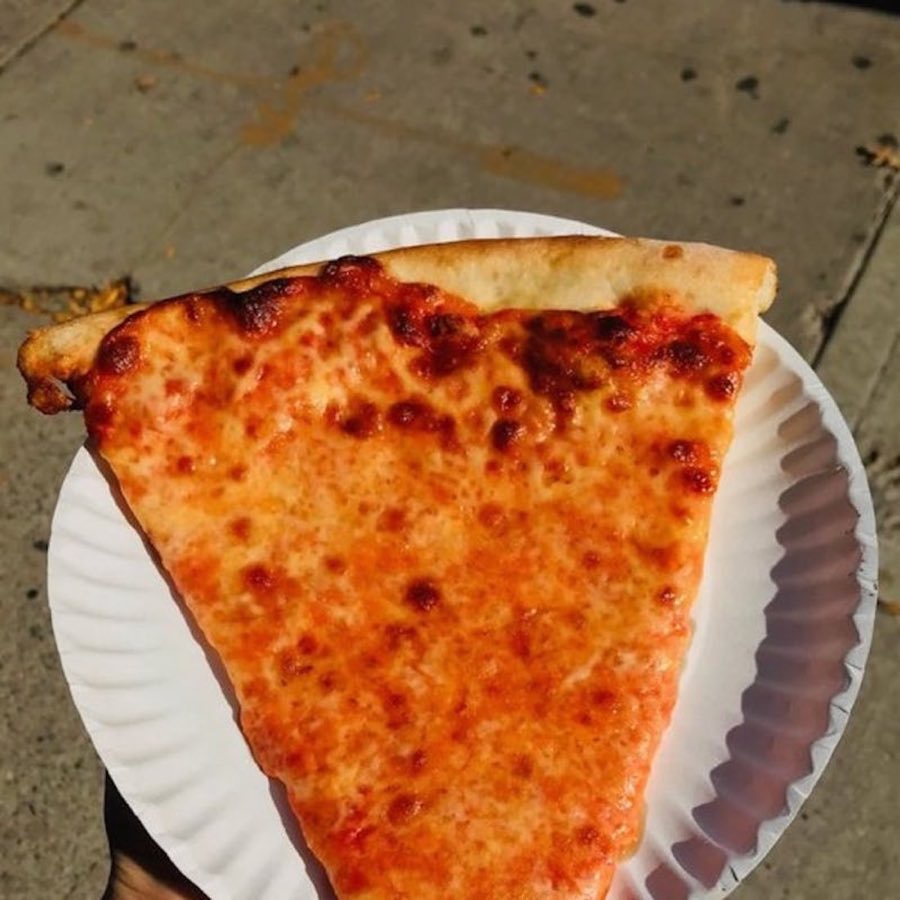 A cheese pizza slice on a paper plate, held out over a New York City sidewalk