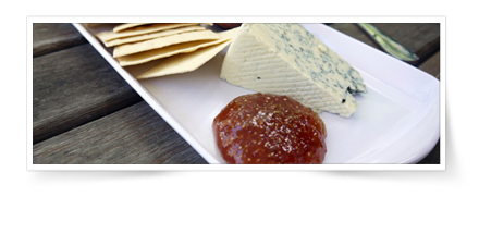 A white plate with a wedge of blue-veined cheese, thin crackers, and a small dish of fruit preserve on a weathered wood table