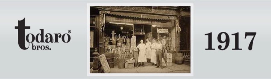 Sepia photograph from 1917 of the original P. Todaro & Co. storefront with a striped awning, olive-oil signage, and three staff in white aprons standing among barrels and crates out front