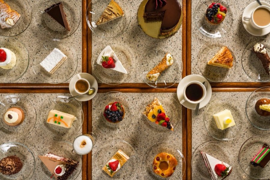 Overhead view of a full Veniero's dessert spread on a marble café table: rainbow cookie, tiramisù, napoleon pastry, fruit tarts, cannoli, mousse cakes, espresso and cappuccino