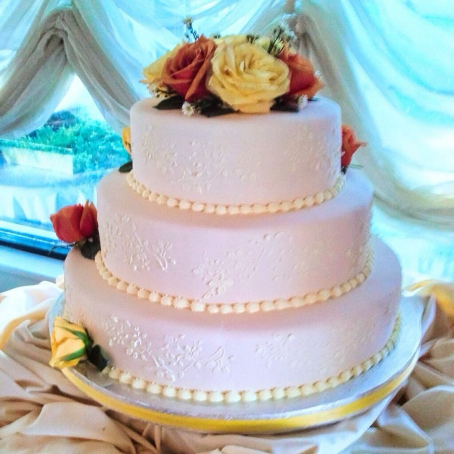 A three-tier pale pink wedding cake with lace-pattern icing, fresh yellow and orange roses between tiers